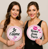 Two young women holding colorful Paris-themed party props with phrases 'Je t'aime' and 'Kisses from Paris'.