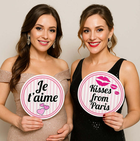 Two young women holding colorful Paris-themed party props with phrases 'Je t'aime' and 'Kisses from Paris'.