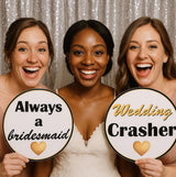 Three women joyfully holding wedding photo booth props, 'Always a bridesmaid' and 'Wedding Crasher,' in front of sparkling backdrop.